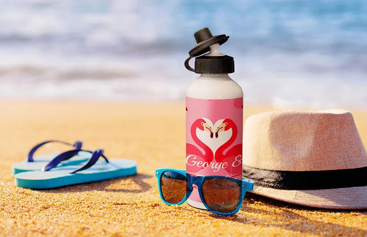 Bottiglia d'acqua personalizzata con fenicottero su spiaggia sabbiosa, accanto a occhiali da sole e cappello. Regalo estivo perfetto!