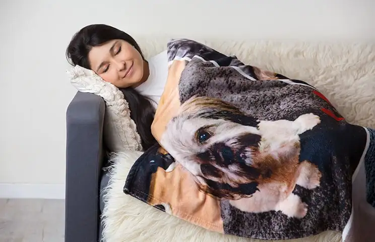 Una donna avvolta in una coperta di vison personalizzata di Printerpix con un design di cani. È felicemente addormentata sul divano.